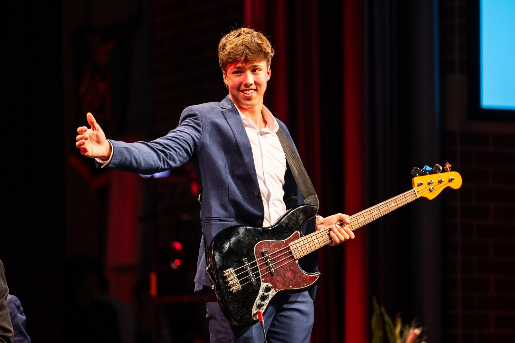 A student centre stage at Soli holding a bass guitar smiling and gesturing to the side of stage