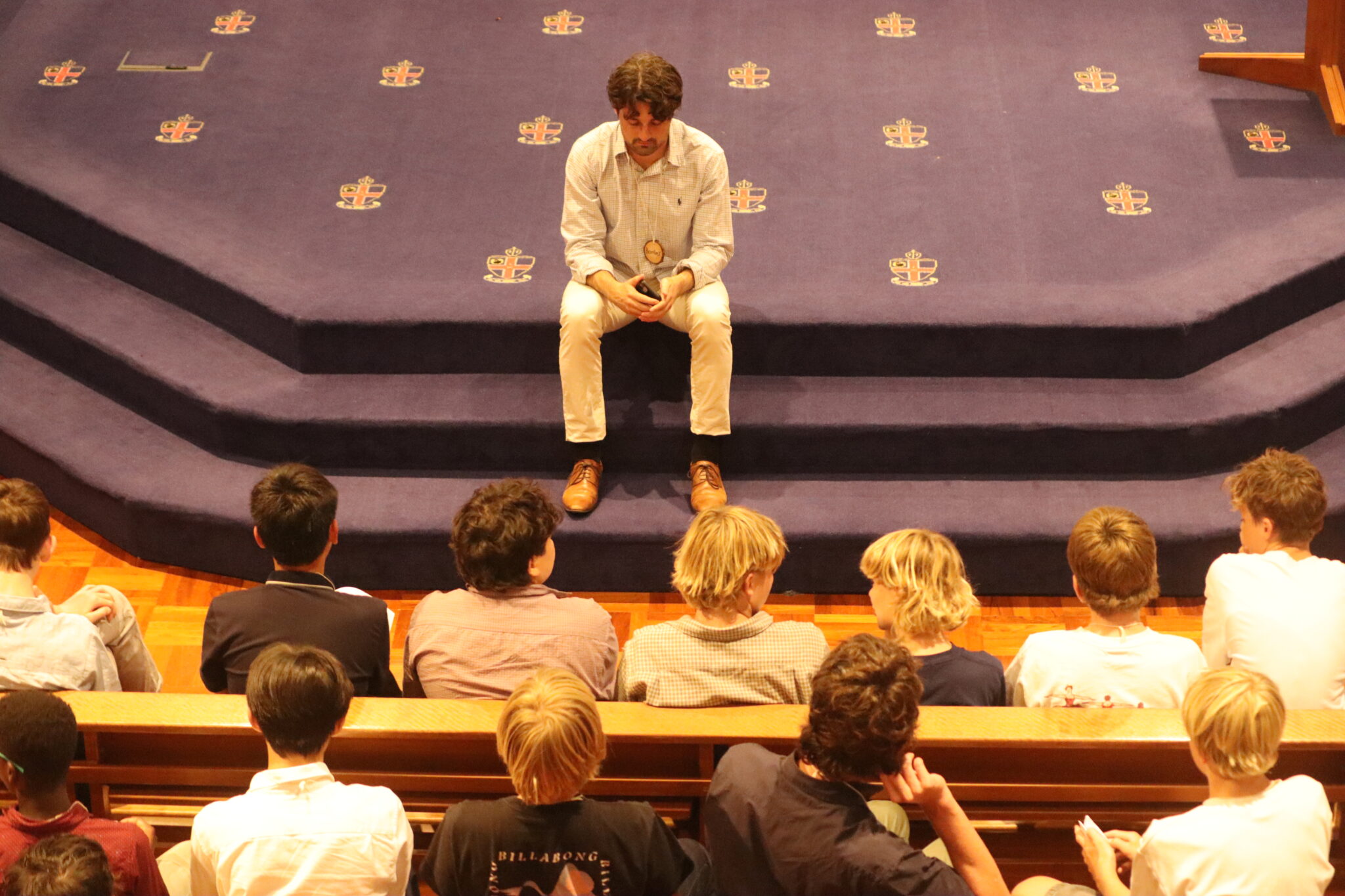 Teacher sits on the Chapel stage in front of students in pews looking reflective